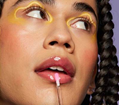 A close-up on a woman’s face. Her makeup includes bright yellow graphic eyeliner, flushed rouge cheeks, and plump lips