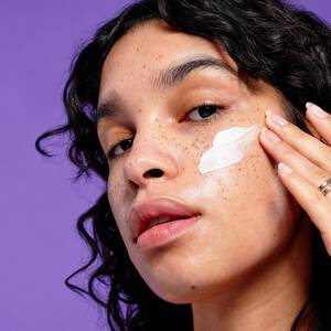 A young woman with a freckled face applies a swipe of NYX Marsh Mellow Primer across her cheek.