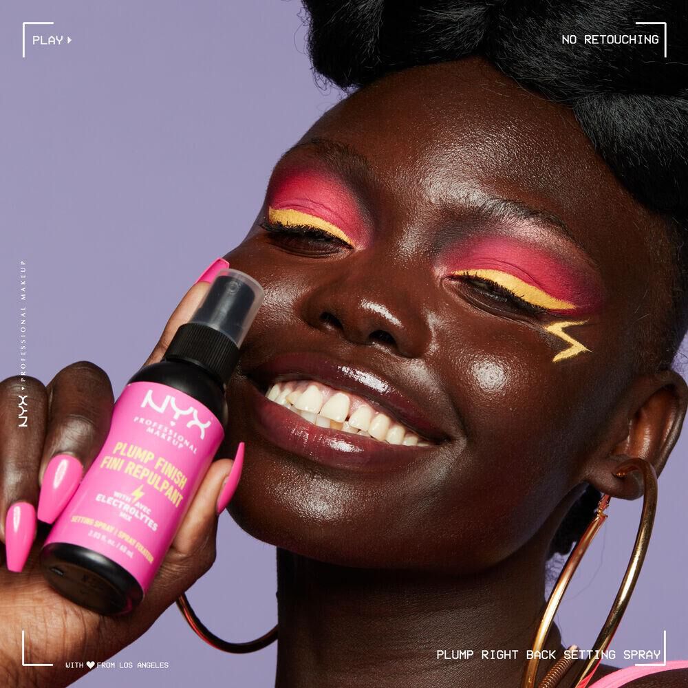 A young woman with bright eye makeup holds a bottle of NYX’s Plump Finish Setting Spray up to her face.