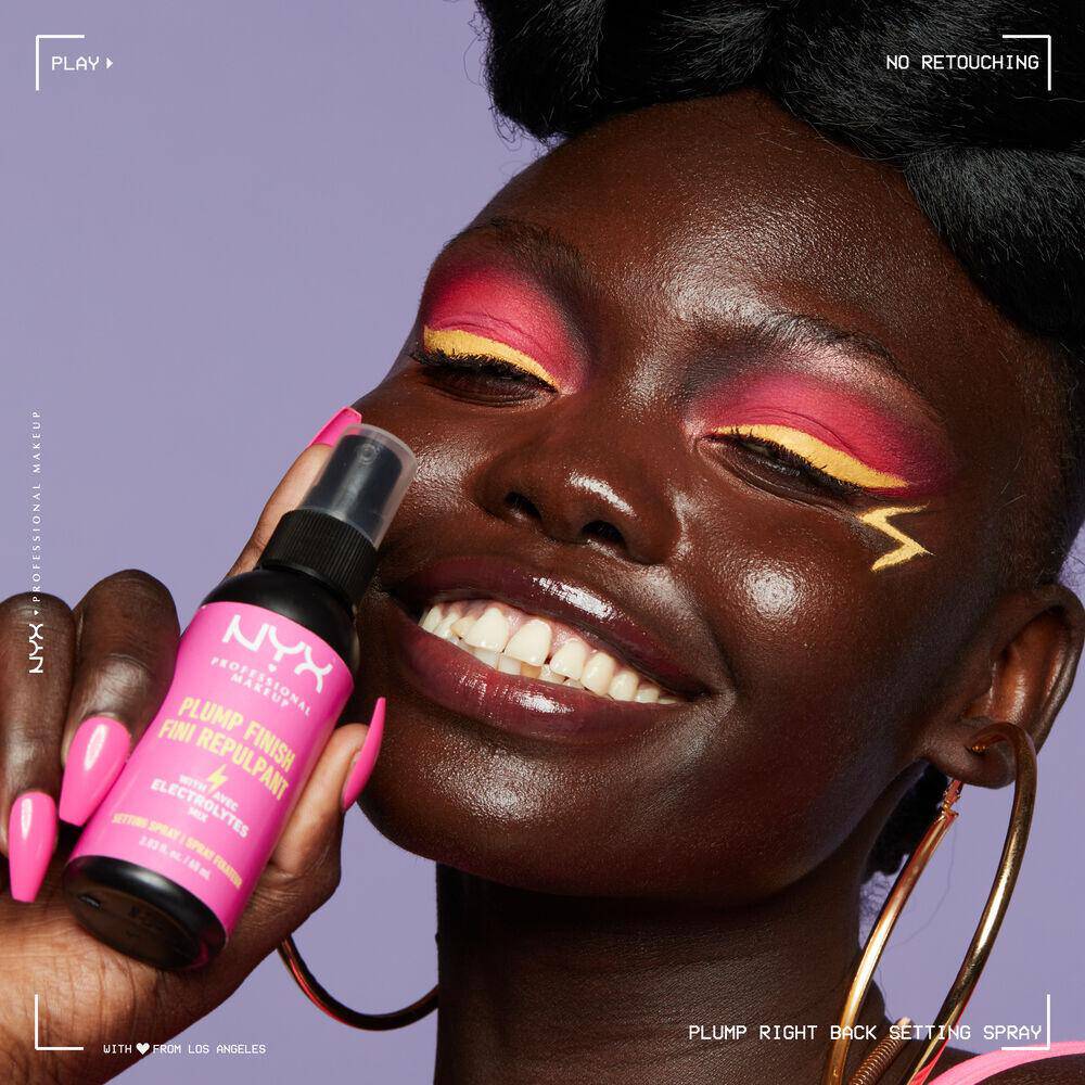 A young woman with bright eye makeup holds a bottle of NYX’s Plump Finish Setting Spray up to her face.